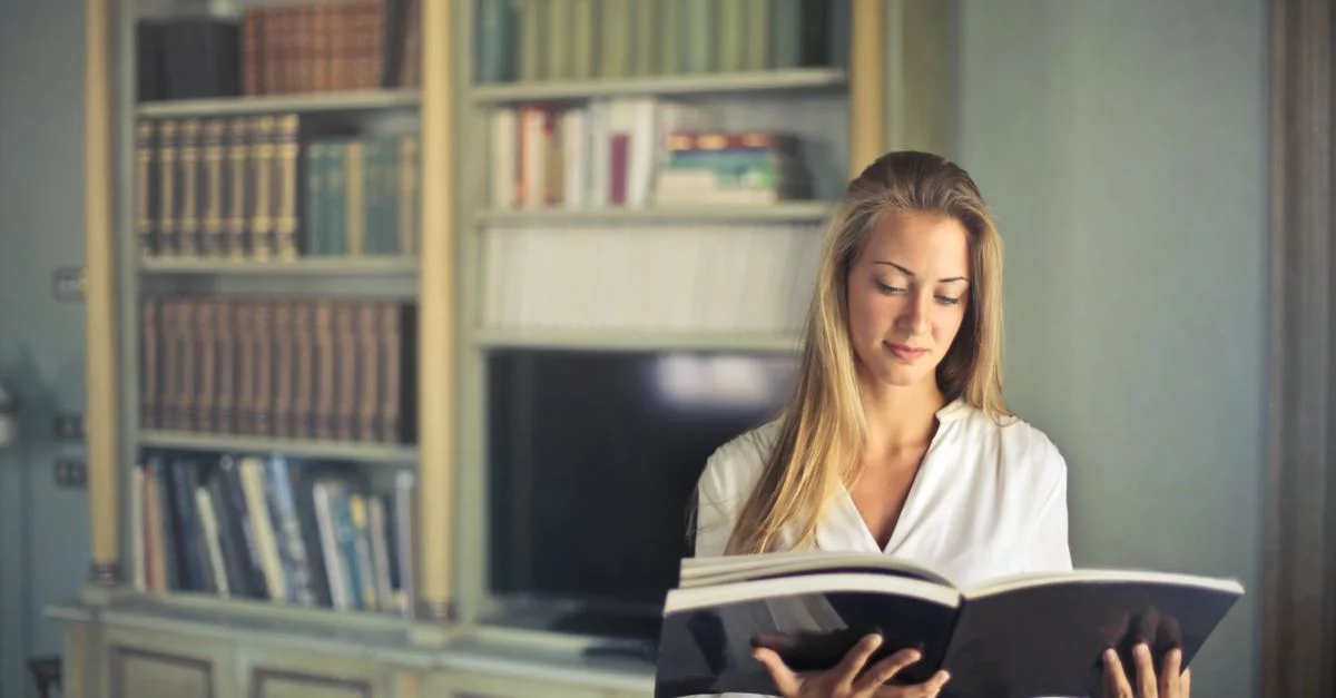 Une femme en haut blanc lisant un livre dans une bibliothèque à domicile, entourée d'étagères.