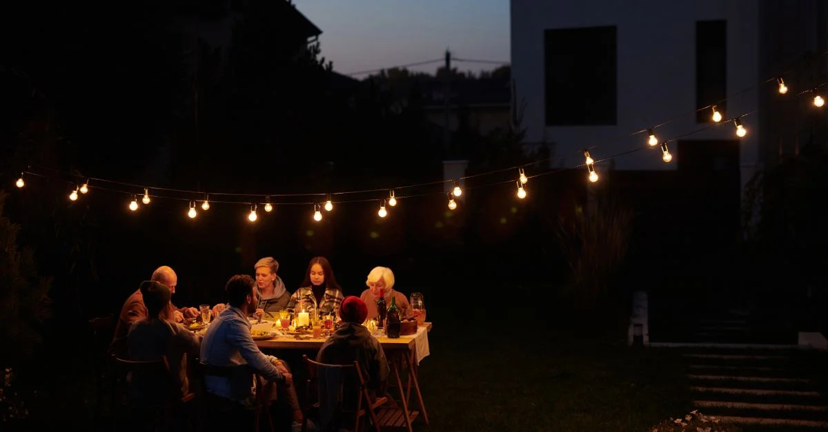 Une famille déguste un repas du soir en plein air sous des guirlandes lumineuses décoratives dans un cadre de jardin.