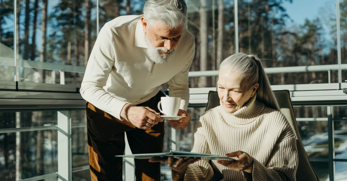 Couple senior appréciant un café et lisant dans une pièce ensoleillée avec de grandes fenêtres.
