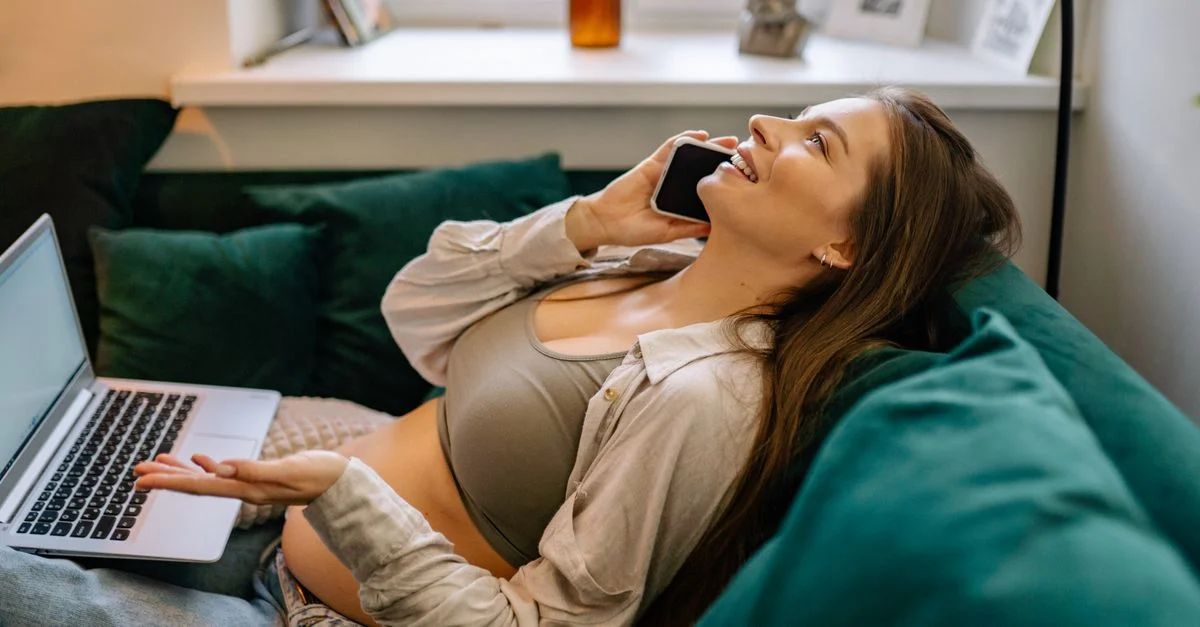 Femme enceinte se relaxant sur un canapé avec un ordinateur portable, profitant d'un appel téléphonique.